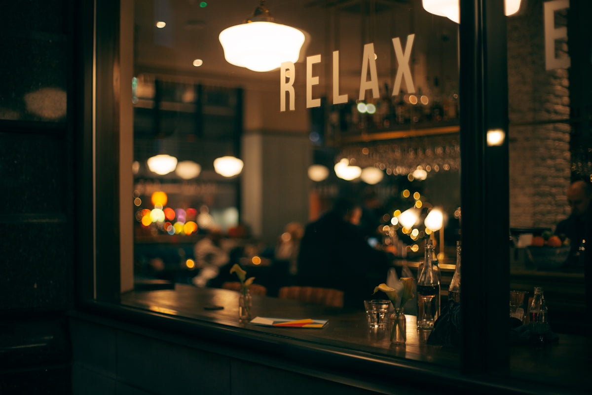 窓ガラスに「RELAX」と書かれたバーやカフェの内部。柔らかな照明とボケた背景が、落ち着いた夜の雰囲気を演出しています。カウンターにはグラスや花瓶が並び、人々がくつろぐ姿がぼんやりと見えます。