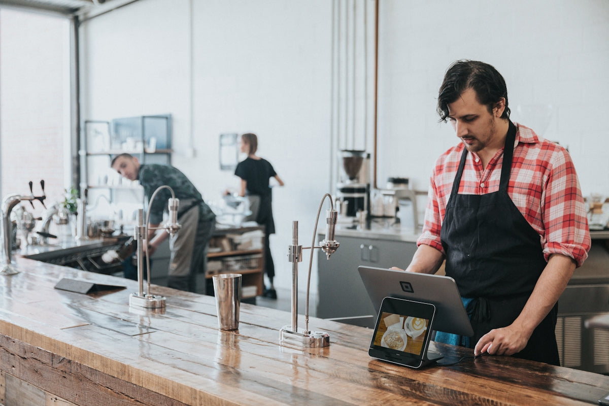 木製カウンターでタブレットを操作するバリスタ。背景には作業中のスタッフと明るいカフェ空間が広がる。