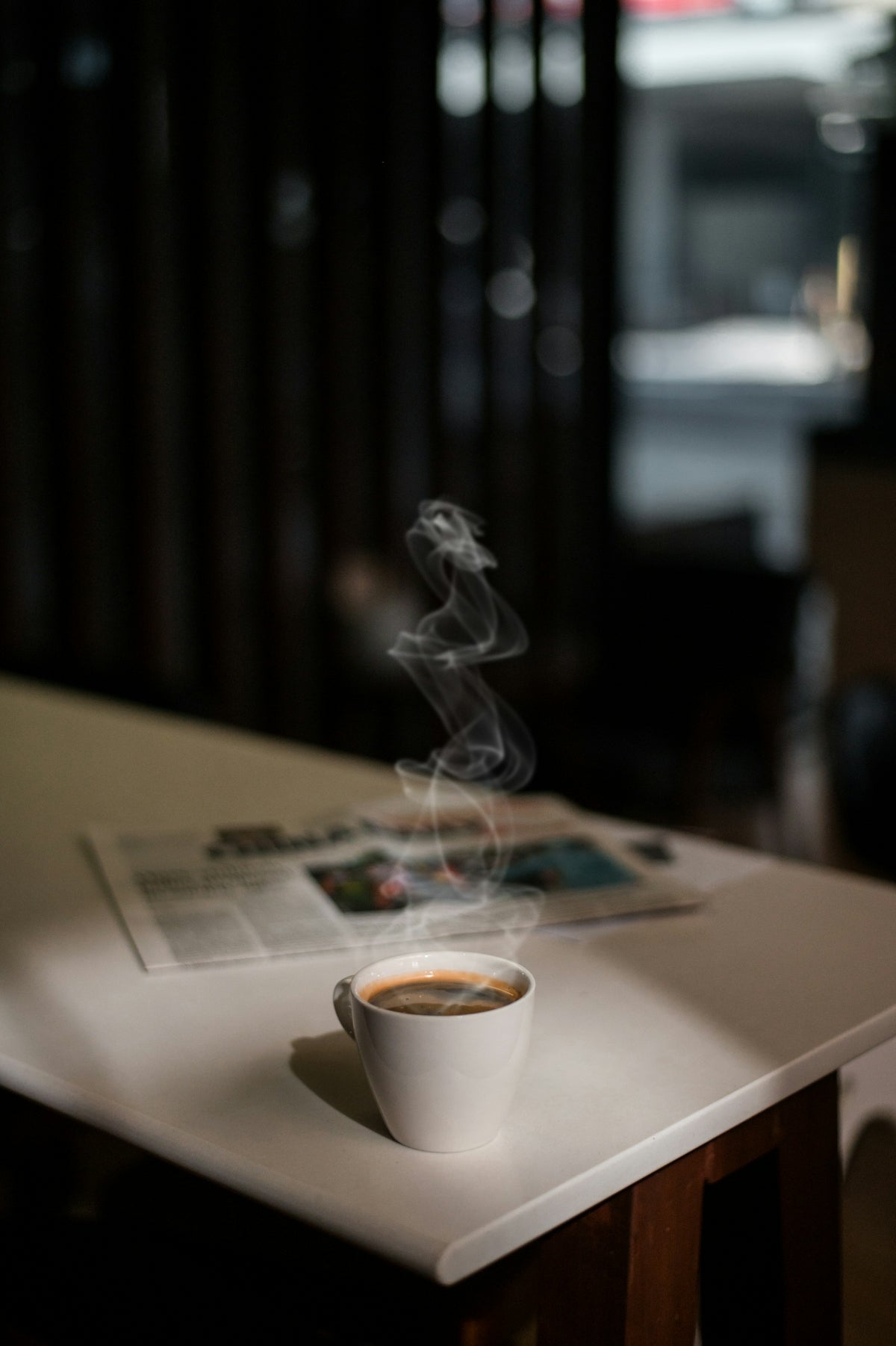 白いコーヒーカップから立ち上る湯気。テーブルには新聞が置かれ、柔らかな光が静かで温かな朝のひとときを演出。