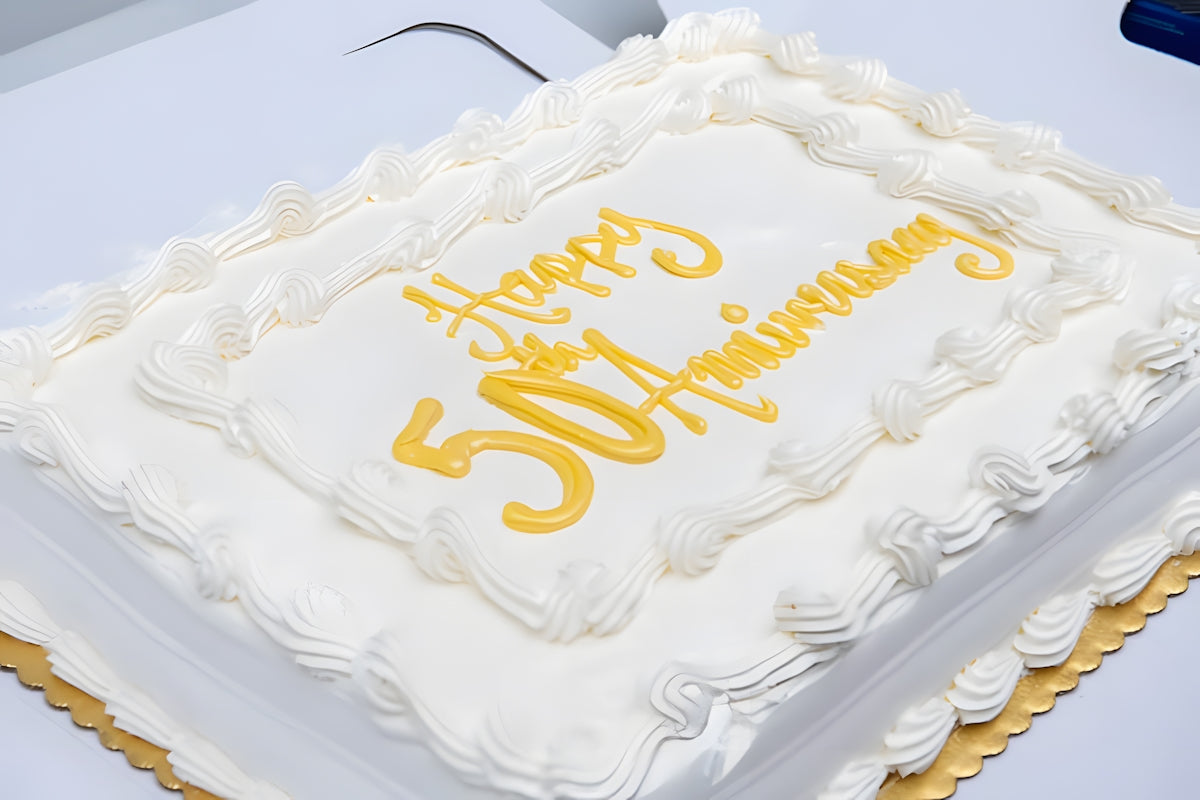 白いホイップで縁取りされた長方形のケーキ。金色の「Happy 50th Anniversary」の文字が輝き、記念すべき50周年を祝う華やかな一品。