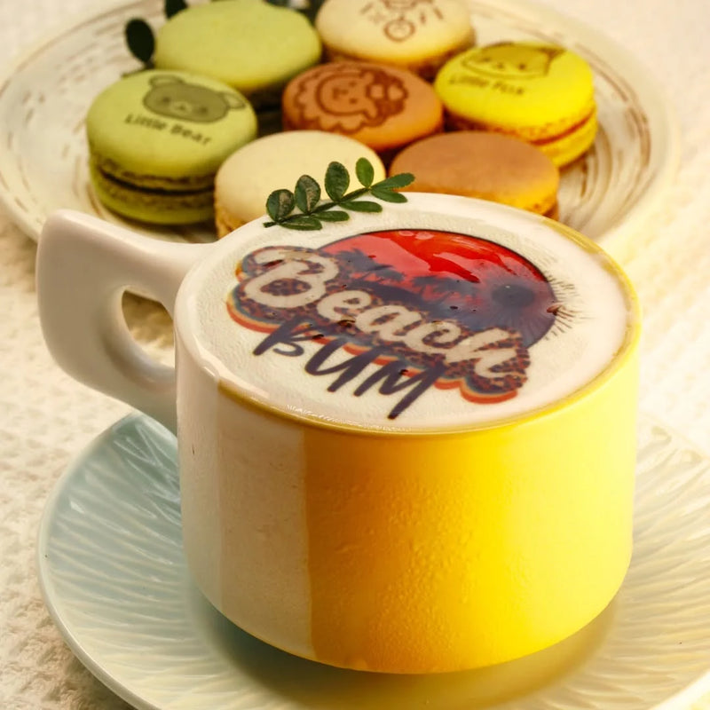 A cup of coffee with a tropical design printed on the foam, with macarons also decorated with edible ink prints.