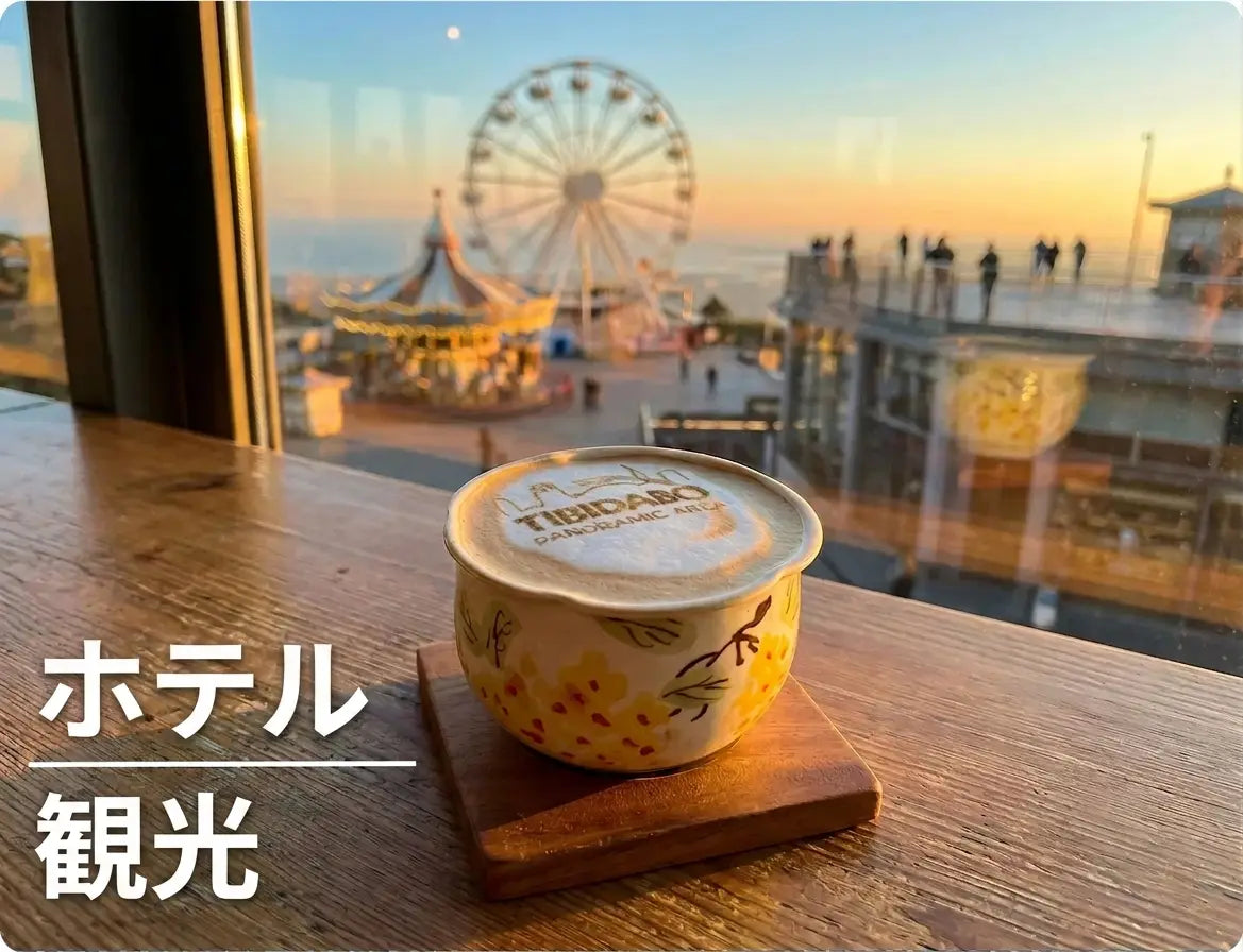 ホテルの窓辺で観光客が楽しむ夕暮れ景色を背景に、コーヒープリンターで「TIBIDABO PANORAMIC AREA」と印刷されたラテアートのコーヒーが置かれた様子。